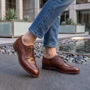 Op de afbeelding: Bruine leren herenschoenen met een klassiek ontwerp. De schoenen hebben een vetersluiting en een gepolijste afwerking. De afbeelding toont de schoenen vanuit een lage hoek, waarbij hun vorm en details worden benadrukt. De schoenen worden gedragen met een blauwe jeans.