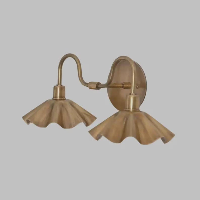 May include: A double wall sconce light fixture with a metallic brass finish. The fixture features two downward-facing, fluted, petal-shaped shades attached to a central horizontal arm that connects to a circular wall-mounting plate.