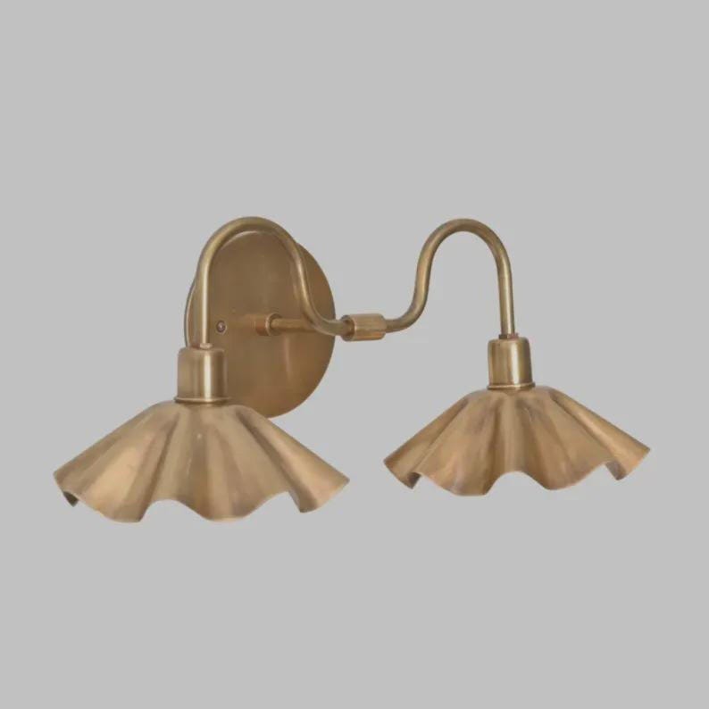 May include: A double wall sconce light fixture with a metallic brass finish. It features a circular wall mounting plate and two curved arms extending to support two downward-facing, fluted, bell-shaped shades with scalloped edges.