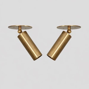 May include: Two brass-colored cylindrical spotlights with a circular base and a ball joint. The spotlights are angled and appear to be mounted on a white surface. The design is modern and minimalist.