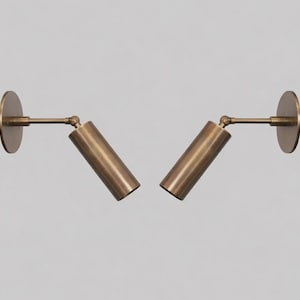 May include: Two brass-toned wall-mounted spotlights. Each spotlight has a circular base, an adjustable arm, and a cylindrical light fixture. The fixtures are angled away from each other, suggesting directional lighting. The design is minimalist and modern.