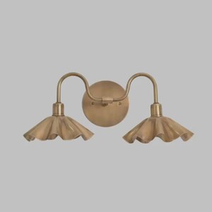 May include: A double wall sconce light fixture with a circular backplate and two curved arms extending outward. Each arm supports a downward-facing, fluted metal shade with a scalloped edge, all finished in a brushed antique brass tone.