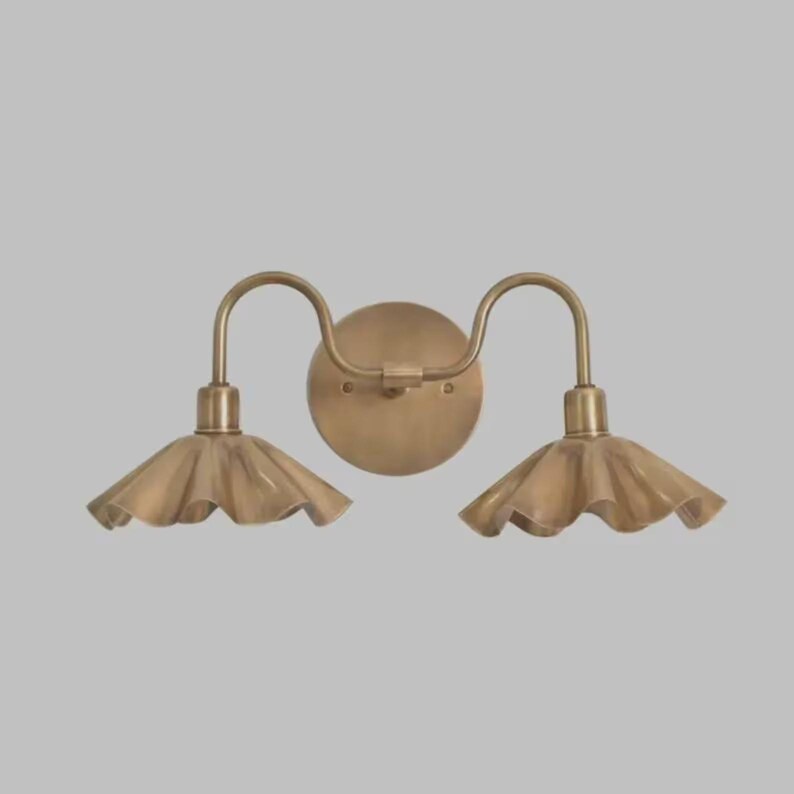 May include: A double wall sconce light fixture with a circular backplate and two curved arms extending outward. Each arm supports a downward-facing, fluted metal shade with a scalloped edge, all finished in a brushed antique brass tone.