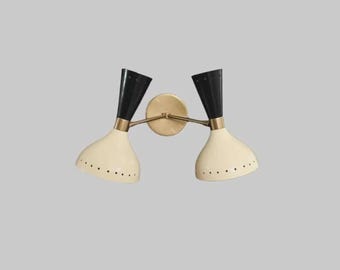 May include: A dual-head wall sconce featuring two cone-shaped shades with black upper sections and cream-colored lower sections. The shades are connected by brass-toned metal arms to a central circular wall-mounting plate in a matching brass finish.