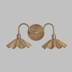 May include: A double wall sconce light fixture with a circular backplate and two curved arms extending outward. Each arm supports a downward-facing, fluted, bell-shaped shade. The entire fixture is finished in a brushed brass metallic tone.