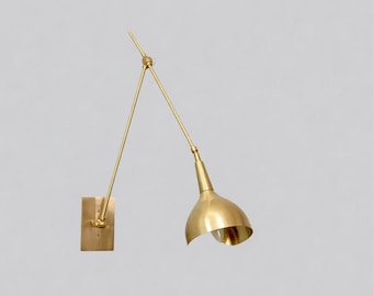 May include: A wall-mounted lamp featuring a brushed brass finish. The fixture consists of a rectangular wall plate, a long adjustable arm with a central pivot point, and a conical shade that directs light downward from a single bulb socket.