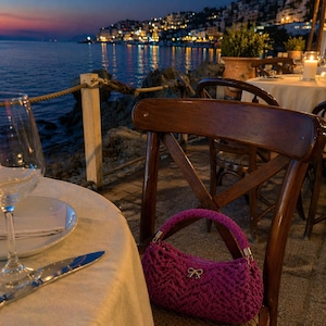 Può includere: Una borsa all'uncinetto fucsia con un dettaglio a fiocco è appoggiata su una sedia di legno in un ristorante all'aperto. La scena è ambientata al tramonto, con vista sul mare e sulle luci della città sullo sfondo. Un tavolo è apparecchiato.