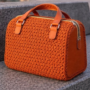 May include: An orange crochet handbag with leather accents. The bag features a textured crochet body, leather handles, and a gold zipper. The bag is a rectangular shape and has a structured design.
