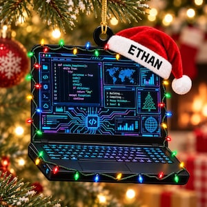 May include: A black laptop-shaped Christmas ornament featuring a screen with code and a Santa hat on the top right corner with the name ETHAN printed on the white brim. The ornament is wrapped in a string of colourful miniature holiday lights.