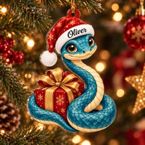 Könnte beinhalten: Ein flacher Weihnachtsbaumschmuck in Form einer blauen Schlange mit großen Augen, die eine rot-weiße Weihnachtsmütze mit dem Namen Oliver auf dem Rand trägt. Die Schlange ist um ein rotes Geschenkpaket mit goldener Schleife gewickelt.