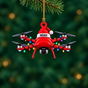 Pode incluir: Um enfeite de Natal em forma de drone vermelho com um gorro de Pai Natal no topo, com o nome NOAH impresso a preto na aba branca. O drone está envolvido por um fio de luzes de Natal coloridas em miniatura e pendurado por um cordão dourado.
