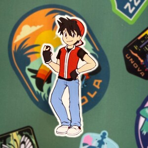 Op de afbeelding: Een sticker met een stripfiguur in een rood vest, zwart shirt en blauwe jeans. De sticker is omgeven door andere stickers met verschillende ontwerpen en tekst, waaronder "Johto", "Unova" en "Kanto". De achtergrond is effen groen.