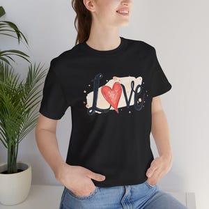 Puede incluir: Camiseta negra con la palabra "Love" en fuente cursiva y un corazón rojo que reemplaza la "o". El diseño incluye un fondo beige estilo acuarela con pequeñas motas marrones. La camiseta está hecha de un material suave.