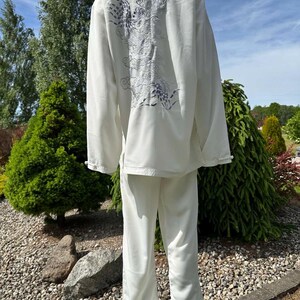 Op de afbeelding: Een wit vechtsportuniform, bestaande uit een top met lange mouwen en een broek. De achterkant van de top heeft een geborduurd drakenontwerp in grijstinten en paars. Het uniform wordt buiten op een mannequin getoond.