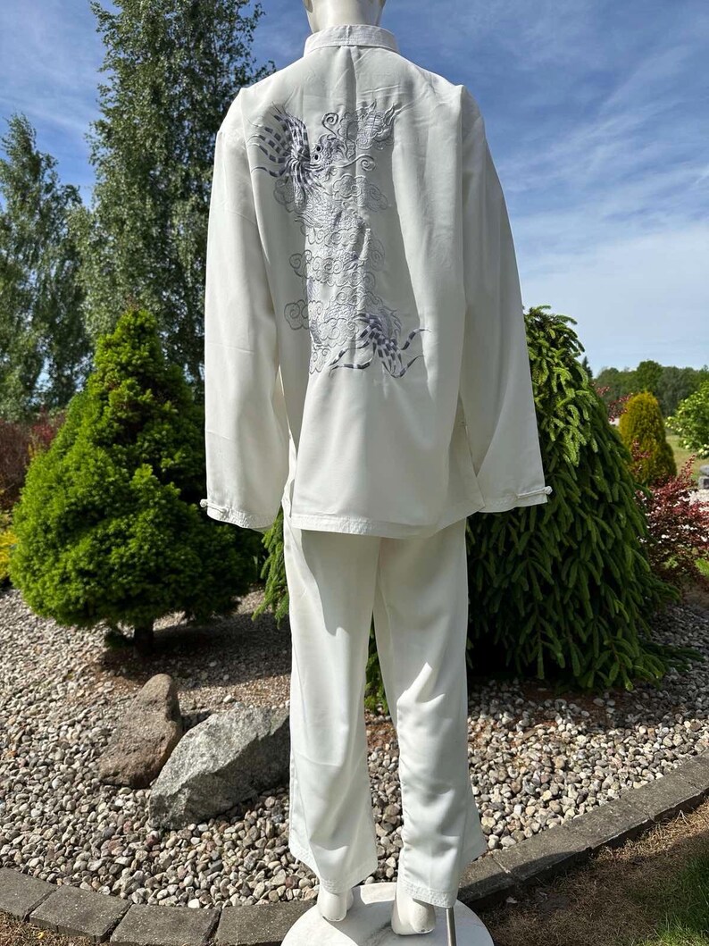 Op de afbeelding: Een wit vechtsportuniform, bestaande uit een top met lange mouwen en een broek. De achterkant van de top heeft een geborduurd drakenontwerp in grijstinten en paars. Het uniform wordt buiten op een mannequin getoond.