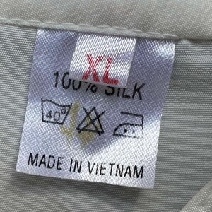 Op de afbeelding: Een close-up van een wit stoffen label met de tekst "XL" in rood, "100% ZIJDE" en wasvoorschriften. Het label vermeldt ook "MADE IN VIETNAM". Het label is bevestigd aan een lichtgrijze stof.