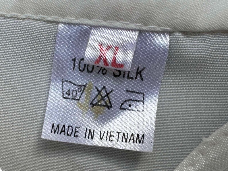 Op de afbeelding: Een close-up van een wit stoffen label met de tekst "XL" in rood, "100% ZIJDE" en wasvoorschriften. Het label vermeldt ook "MADE IN VIETNAM". Het label is bevestigd aan een lichtgrijze stof.
