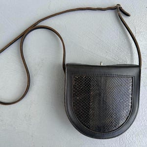 May include: Black leather crossbody bag with a brown snakeskin panel on the front. The bag has a brown leather strap and a silver clasp. The bag is a small, vintage style.