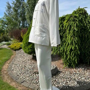 Op de afbeelding: Een witte tweedelige outfit met een jas met lange mouwen, kikkerknopen en geborduurde details, gecombineerd met een bijpassende broek met wijde pijpen. De set wordt buiten op een mannequin getoond en toont een klassieke, elegante stijl.