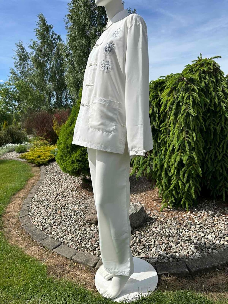 Op de afbeelding: Een witte tweedelige outfit met een jas met lange mouwen, kikkerknopen en geborduurde details, gecombineerd met een bijpassende broek met wijde pijpen. De set wordt buiten op een mannequin getoond en toont een klassieke, elegante stijl.