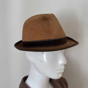 Könnte beinhalten: Ein beigefarbener Fedora-Hut mit einer glatten, matten Oberfläche und einem dunkelbraunen Samtband um die Basis der Krone. Der Hut verfügt über eine strukturierte Krempe mit einem dunkelbraunen Rand, der für eine klare Formgebung sorgt.