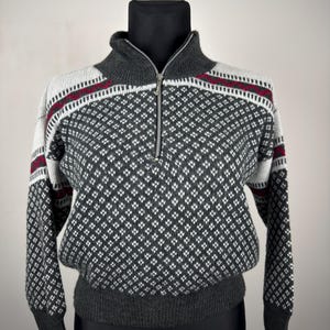 May include: A gray, white, and burgundy patterned sweater with a high collar and a quarter-zip closure. The sweater features a diamond pattern in gray and white, with burgundy stripes on the shoulders and sleeves. The sweater is displayed on a black mannequin.