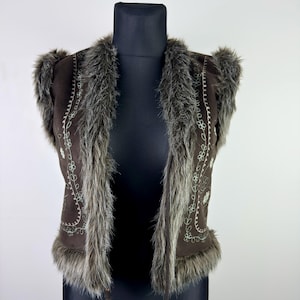 Könnte beinhalten: Eine braune Wildlederweste mit Kunstfellbesatz und gestickten floralen Details. Die Weste wird auf einer schwarzen Schaufensterpuppe präsentiert. Das Kunstfell ist eine Mischung aus Grau und Braun. Die Weste hat einen Kordelverschluss.