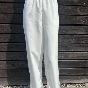 Op de afbeelding: Witte broek met elastische tailleband. De broek is gemaakt van een lichte stof en heeft een losse, vloeiende pasvorm. De broek wordt gepresenteerd op een mannequin tegen een donkere houten achtergrond.