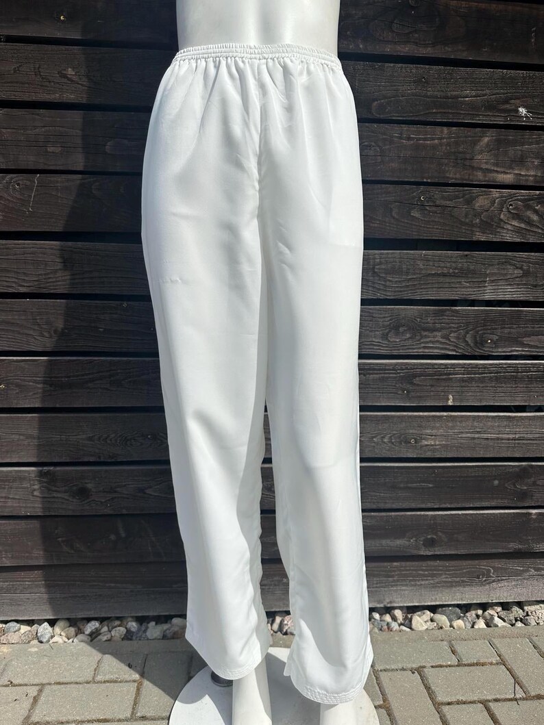 Op de afbeelding: Witte broek met elastische tailleband. De broek is gemaakt van een lichte stof en heeft een losse, vloeiende pasvorm. De broek wordt gepresenteerd op een mannequin tegen een donkere houten achtergrond.