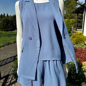 Puede incluir: Un conjunto de tres piezas en azul claro que consta de un blazer sin mangas, un top de cuello cuadrado y unos pantalones cortos. El blazer tiene dos botones y un corte entallado. Los pantalones cortos tienen cintura fruncida. El conjunto se muestra en un maniquí.