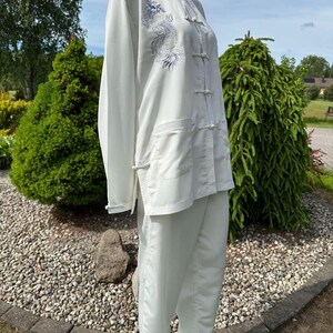 Op de afbeelding: Een wit vechtsportuniform, bestaande uit een top met lange mouwen en een broek, wordt op een mannequin getoond. De top heeft een drakenborduurwerk op de voorkant. De outfit heeft een mandarijnkraag en kikker sluitingen. De broek is losvallend.