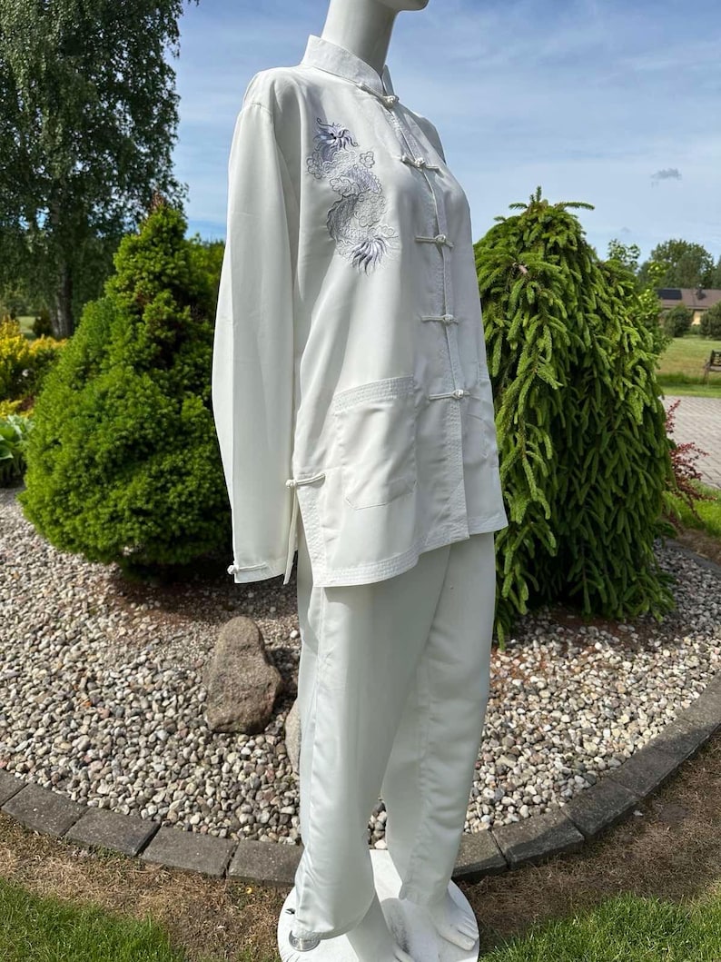 Op de afbeelding: Een wit vechtsportuniform, bestaande uit een top met lange mouwen en een broek, wordt op een mannequin getoond. De top heeft een drakenborduurwerk op de voorkant. De outfit heeft een mandarijnkraag en kikker sluitingen. De broek is losvallend.
