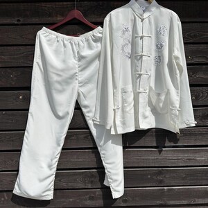 Op de afbeelding: Een witte zijden pyjamaset, bestaande uit een top met lange mouwen en een broek, hangt tegen een donkere houten achtergrond. De top heeft een Chinese kraag, kikkerknopen en geborduurde bloemen- en drakenontwerpen.
