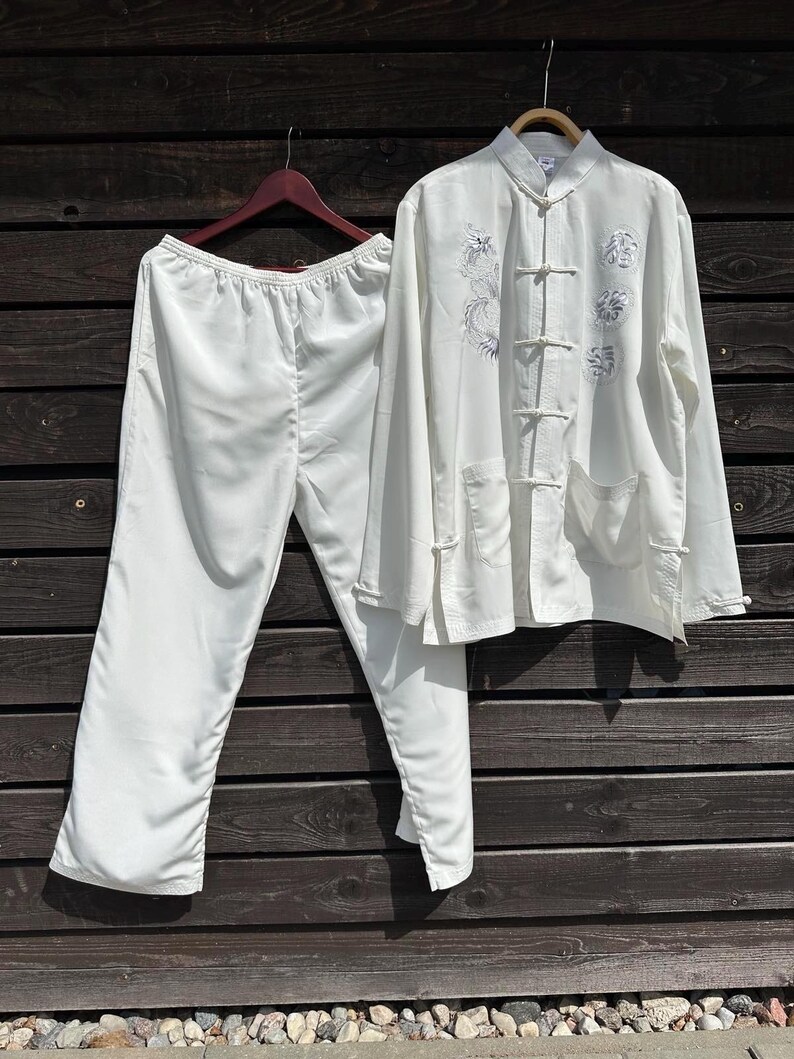 Op de afbeelding: Een witte zijden pyjamaset, bestaande uit een top met lange mouwen en een broek, hangt tegen een donkere houten achtergrond. De top heeft een Chinese kraag, kikkerknopen en geborduurde bloemen- en drakenontwerpen.