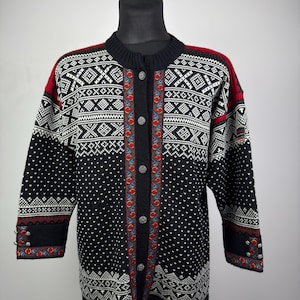 Puede incluir: Cárdigan con estampado negro, blanco y rojo con cierre de botones. El suéter presenta un diseño geométrico con detalles florales y se exhibe en un maniquí negro. Las mangas son de tres cuartos.