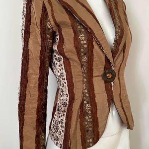 Könnte beinhalten: Braun gestreifter Blazer mit floralen Akzenten. Der Blazer hat einen einzelnen Knopfverschluss und ein fallendes Revers. Die Ärmel sind lang und der Stoff hat eine strukturierte Optik. Der Blazer ist neutralfarben.