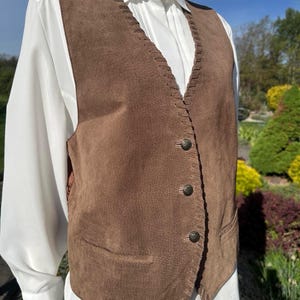 May include: Brown suede vest with decorative stitching and three buttons. The vest has two front pockets and is worn over a white collared shirt. A classic piece of apparel.