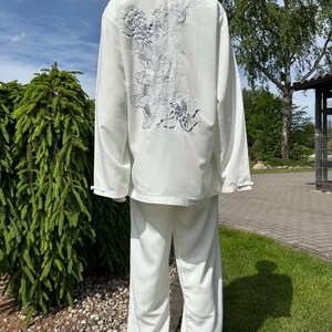 Op de afbeelding: Wit vechtsportuniform, bestaande uit een top met lange mouwen en een broek. De achterkant van de top heeft een zilverkleurig geborduurd drakenontwerp. De outfit wordt buiten op een mannequin getoond, met groen en een blauwe lucht.