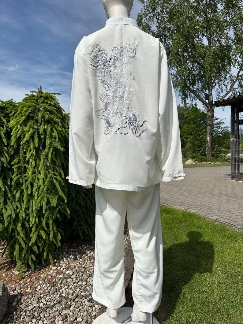 Op de afbeelding: Wit vechtsportuniform, bestaande uit een top met lange mouwen en een broek. De achterkant van de top heeft een zilverkleurig geborduurd drakenontwerp. De outfit wordt buiten op een mannequin getoond, met groen en een blauwe lucht.