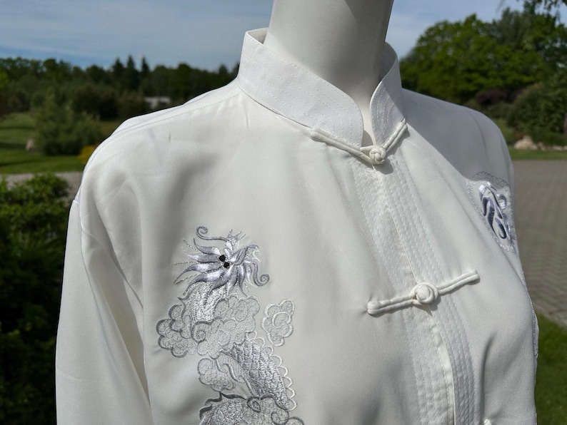 Op de afbeelding: Wit vechtsportuniform met een Chinese kraag en kikker sluitingen. Het uniform heeft ingewikkelde zilveren drakenborduurwerk op de voorkant en schouder. De top met lange mouwen is gemaakt van een gladde, lichte stof.