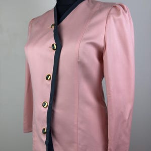 Op de afbeelding: Een vintage roze blazer met zwarte bies en gouden knopen. De blazer heeft lange mouwen en een getailleerde pasvorm. De zwarte bies omlijnt de kraag, voorkant en manchetten. De gouden knopen voegen elegantie toe.
