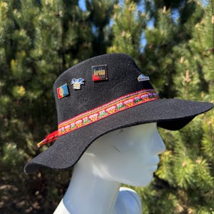 May include: Black wool hat with a wide brim, adorned with colorful pins and a patterned band. The hat features a red and patterned ribbon around the crown. The hat is displayed on a white mannequin head.