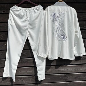 Op de afbeelding: Een witte tweedelige pyjamaset hangt tegen een houten achtergrond. De top heeft een mandarijnkraag en lange mouwen, met een geborduurd drakenontwerp op de rug. De broek heeft rechte pijpen met een elastische tailleband.