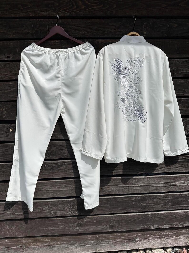 Op de afbeelding: Een witte tweedelige pyjamaset hangt tegen een houten achtergrond. De top heeft een mandarijnkraag en lange mouwen, met een geborduurd drakenontwerp op de rug. De broek heeft rechte pijpen met een elastische tailleband.
