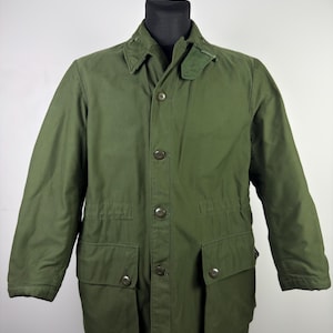 May include: Olive green military-style jacket with a button-up front, collar, and two large front pockets. The jacket has a classic design with a tailored fit, suitable for various weather conditions. The jacket is displayed on a black mannequin.