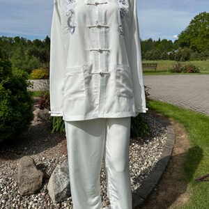 Op de afbeelding: Witte vechtsportuniform met een opstaande kraag, lange mouwen en bijpassende broek. De top heeft een drakenborduurwerk in blauw- en grijstinten. Het uniform heeft een relaxte pasvorm en wordt op een mannequin gepresenteerd.