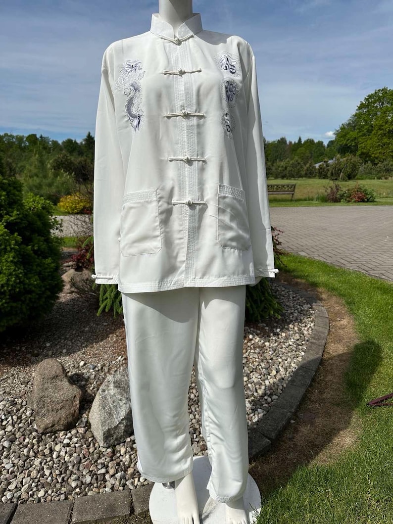 Op de afbeelding: Witte vechtsportuniform met een opstaande kraag, lange mouwen en bijpassende broek. De top heeft een drakenborduurwerk in blauw- en grijstinten. Het uniform heeft een relaxte pasvorm en wordt op een mannequin gepresenteerd.
