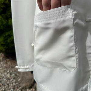 Op de afbeelding: Close-up van een wit vechtsportuniform, met een zakdetail. De zak is rechthoekig met een versterkte bovenrand. De stof lijkt glad en licht reflecterend. Het uniform is schoon en fris.