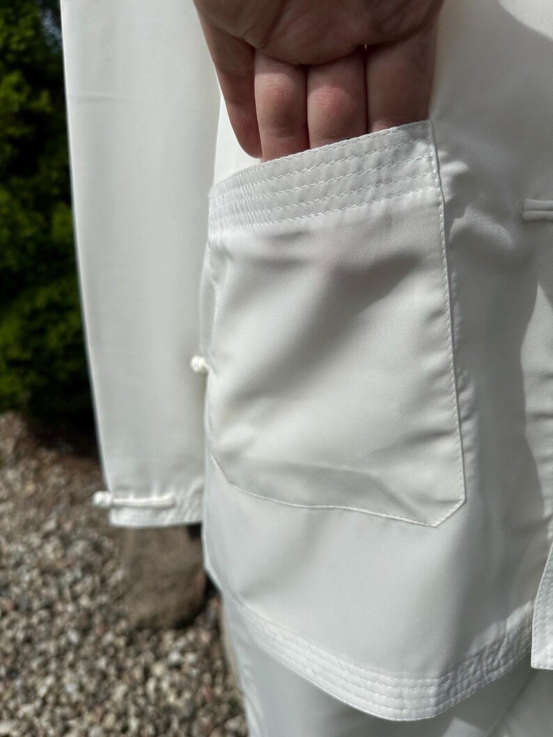 Op de afbeelding: Close-up van een wit vechtsportuniform, met een zakdetail. De zak is rechthoekig met een versterkte bovenrand. De stof lijkt glad en licht reflecterend. Het uniform is schoon en fris.