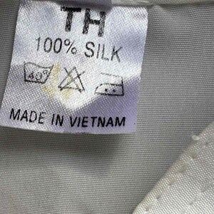 Op de afbeelding: Een wit stoffen label met de tekst "TH", "100% SILK" en "MADE IN VIETNAM" in het zwart. Het label bevat ook wassymbolen. Het label is op een lichtgrijze stof genaaid.
